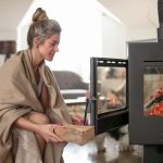 Une femme ajoute une bûche dans un poêle en fonte ouvert, préparant le foyer pour l'hiver.