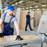 Artisan d'une entreprise de menuiserie découpant avec précision un panneau de bois sur une scie circulaire industrielle dans un grand atelier lumineux.
