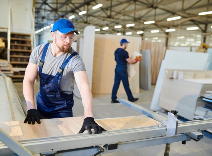 Artisan d'une entreprise de menuiserie découpant avec précision un panneau de bois sur une scie circulaire industrielle dans un grand atelier lumineux.